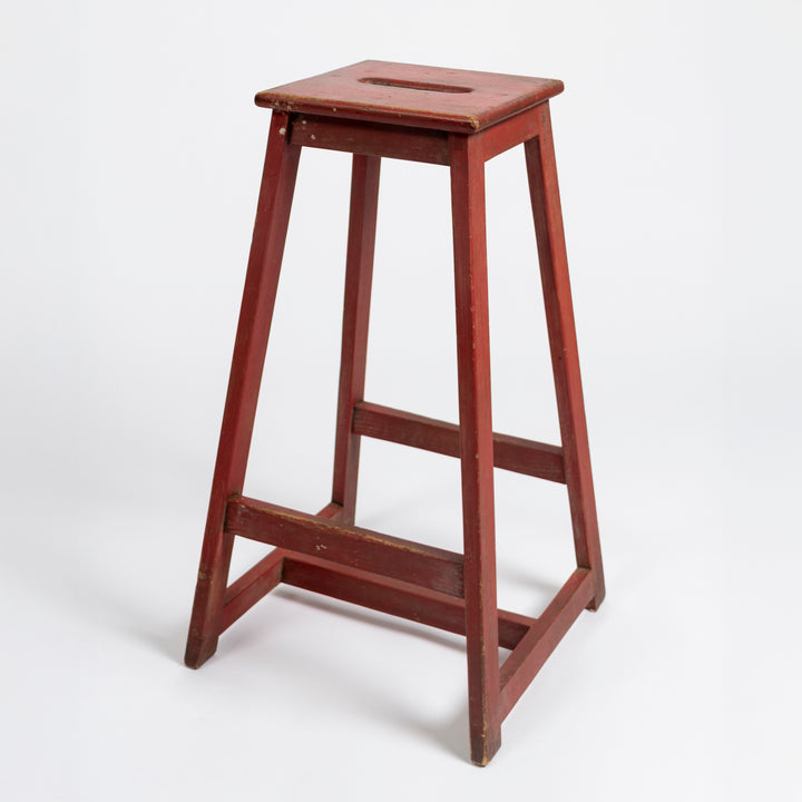 A vintage wooden stool with a reddish-brown painted finish, featuring a square seat with a central cutout and a footrest