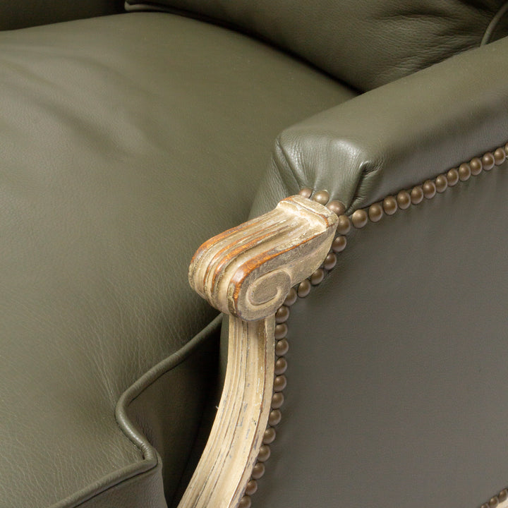 Close-up detail of an upholstered armchair corner showing dark olive green leather, brass nailhead trim, and a distressed carved wooden armrest.