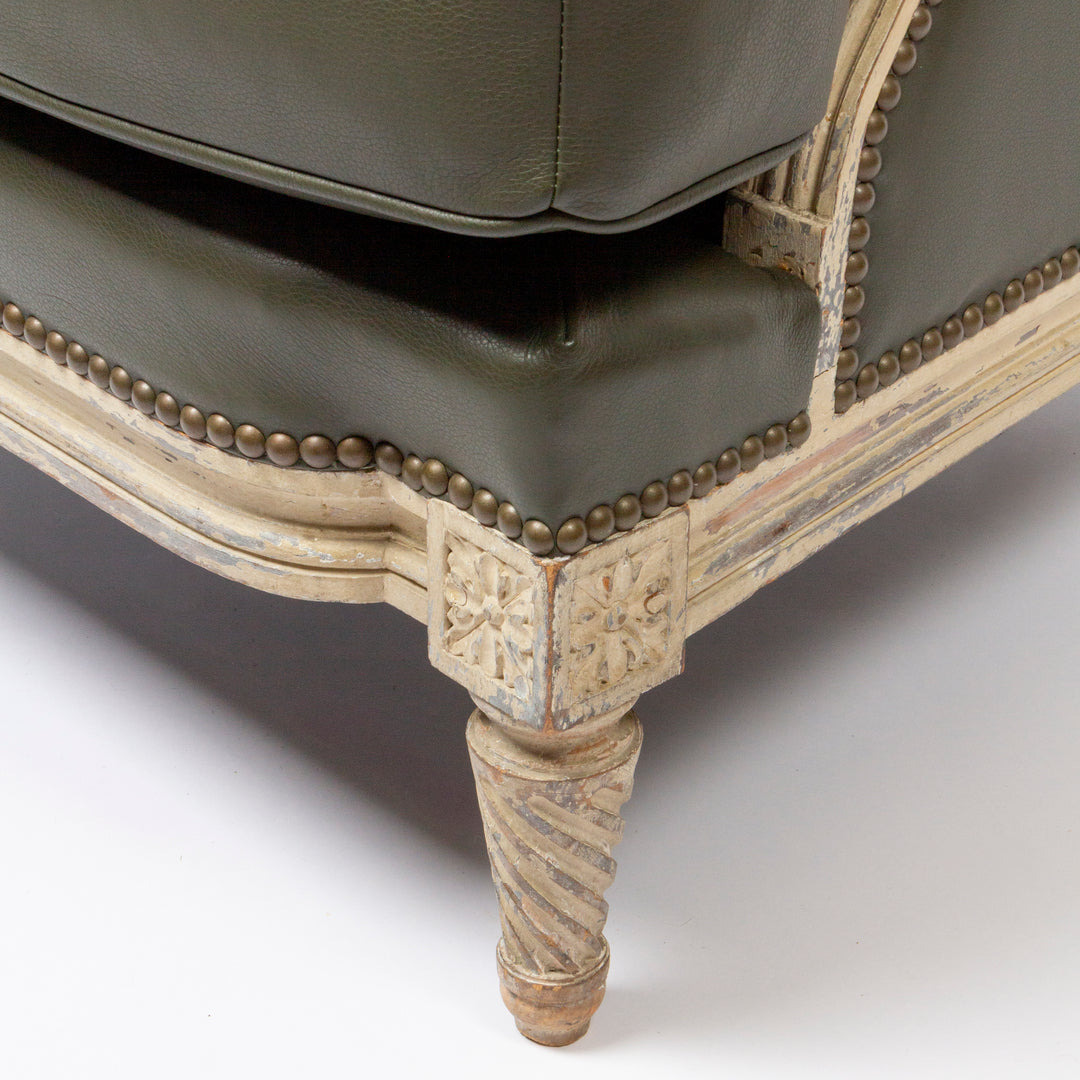 Close-up of an upholstered chair corner showing dark olive green leather seat and back cushions with brass nailhead trim and a distressed cream carved wooden frame on a white studio background.