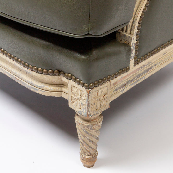 Close-up of an upholstered chair corner showing dark olive green leather seat and back cushions with brass nailhead trim and a distressed cream carved wooden frame on a white studio background.