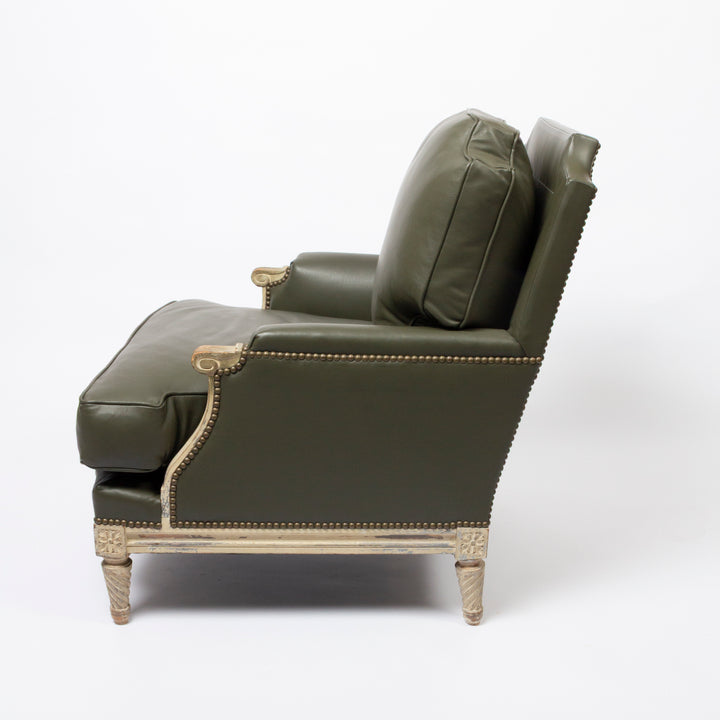 An olive green leather armchair with gold-toned nailhead trim and carved wooden legs, shown from a side profile against a white studio background.