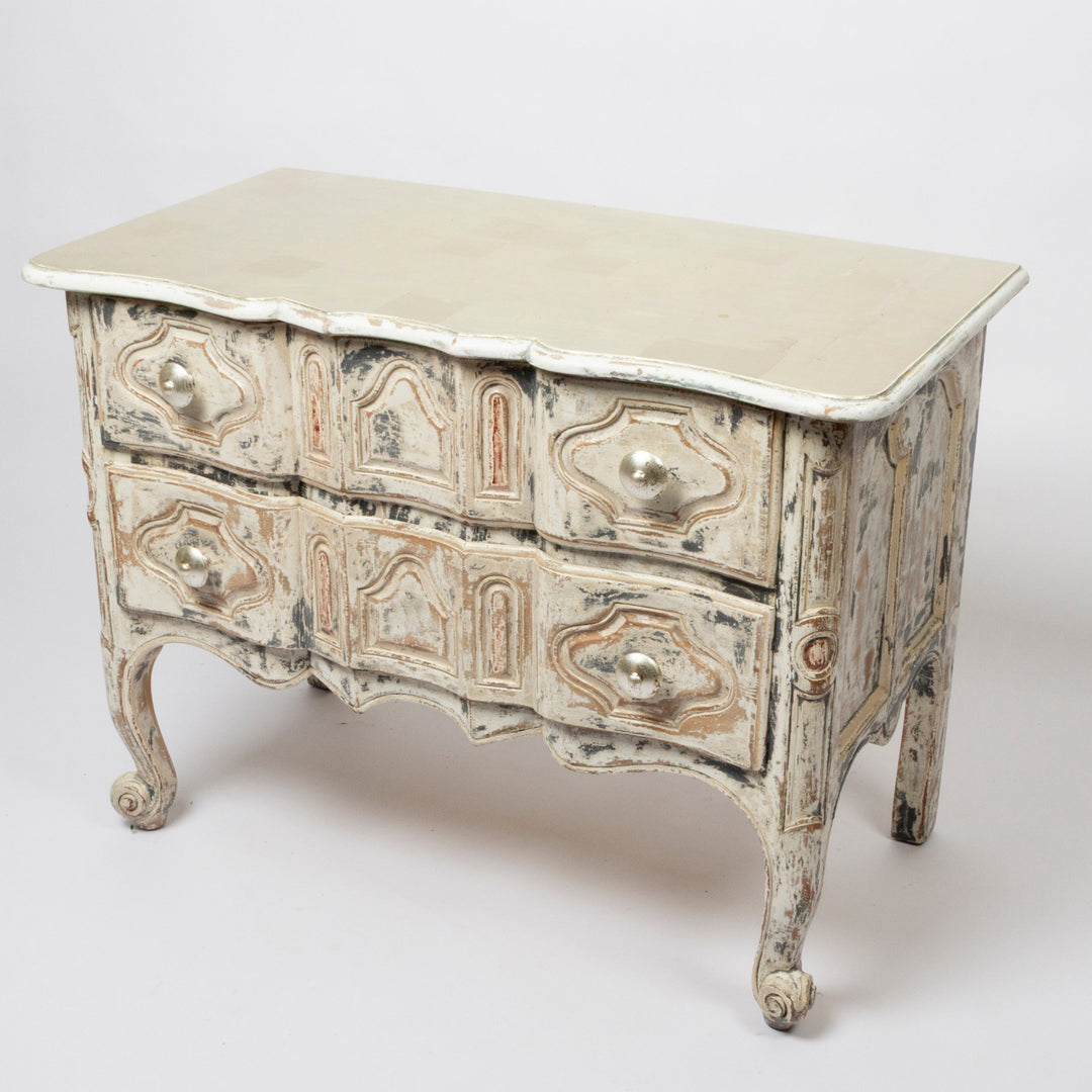 An antique French-style chest of drawers with a distressed finish, featuring a light beige top, carved drawer fronts, and cabriole legs, photographed on a seamless light gray background.