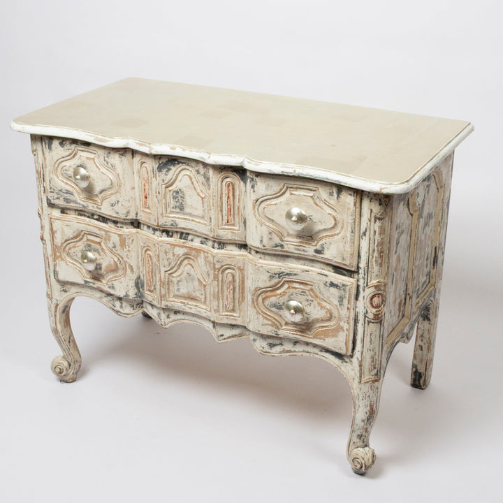 An antique French-style chest of drawers with a distressed finish, featuring a light beige top, carved drawer fronts, and cabriole legs, photographed on a seamless light gray background.
