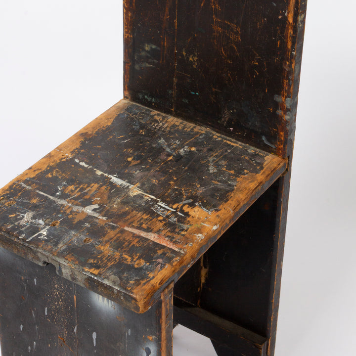A close-up of a distressed wooden chair seat and backrest showing a dark black finish with heavy weathering, peeling paint, and exposed warm brown wood underneath, set against a plain white background.