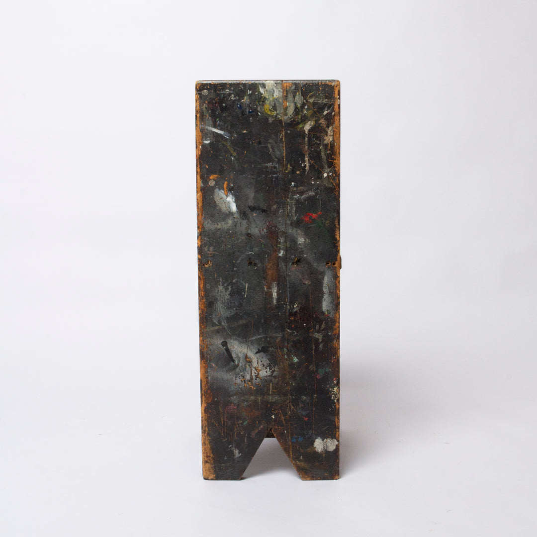 A rectangular dark wooden block or panel with a V-shaped notch at the bottom, showing a distressed black and brown surface with paint-like markings, photographed against a plain light gray background.