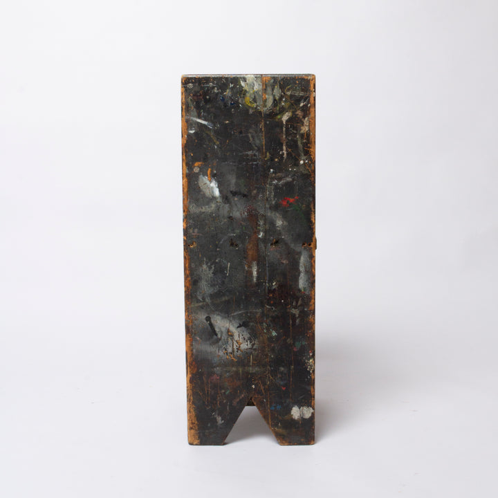 A rectangular dark wooden block or panel with a V-shaped notch at the bottom, showing a distressed black and brown surface with paint-like markings, photographed against a plain light gray background.