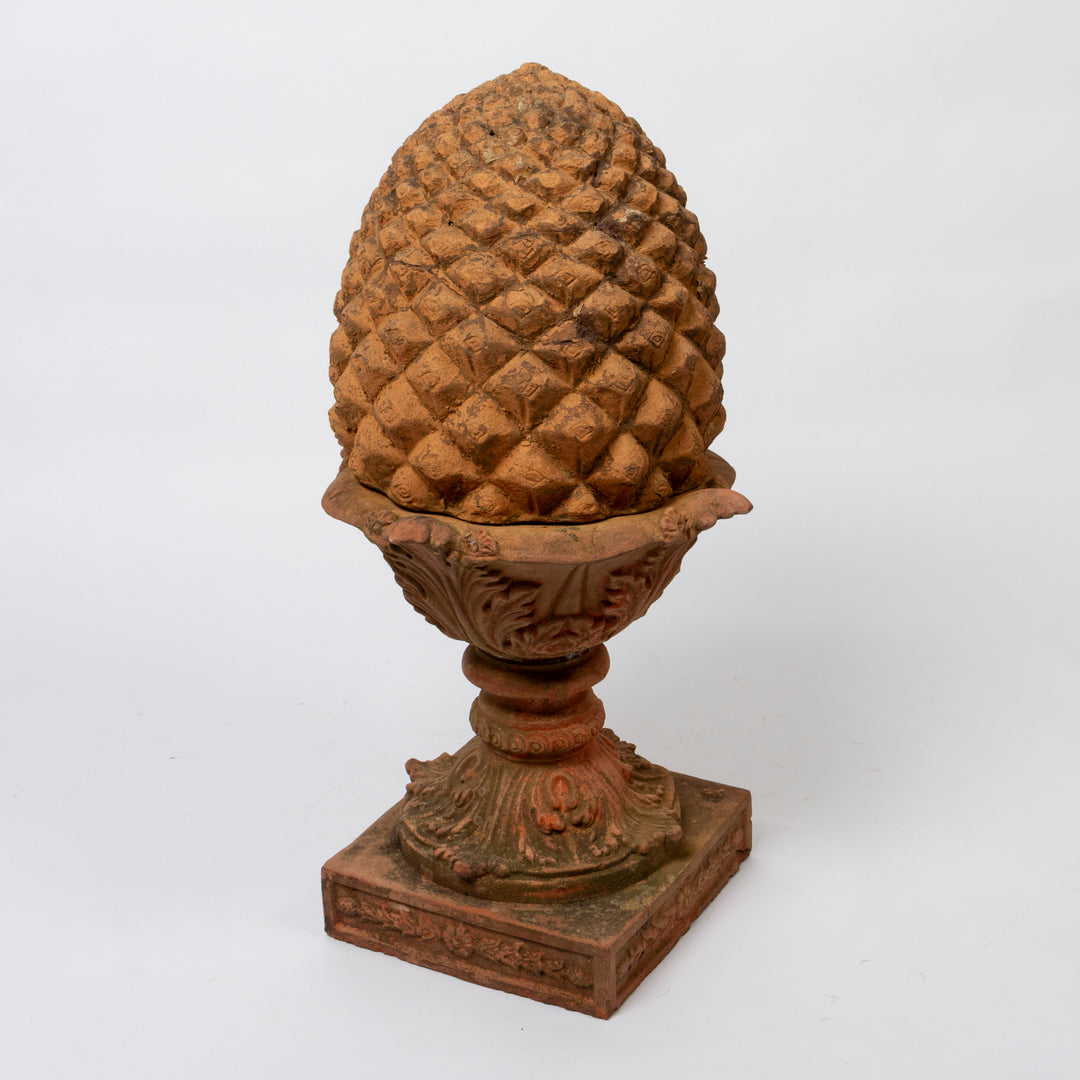 A decorative terracotta garden ornament shaped like a pinecone resting on an ornate pedestal base, photographed against a plain white background.