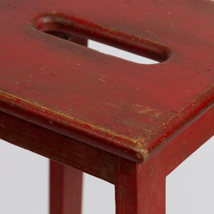 Close-up of a distressed red wooden stool or table corner with a cutout handle on the seat, showing worn paint and natural wood showing through.
