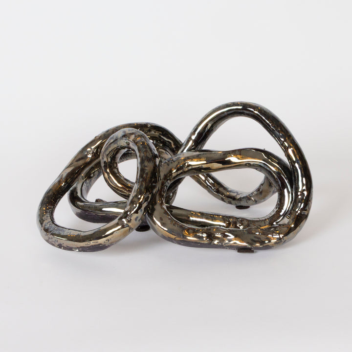 An abstract sculptural object made of interlocking glossy loops with a metallic dark finish, photographed on a clean white studio background.