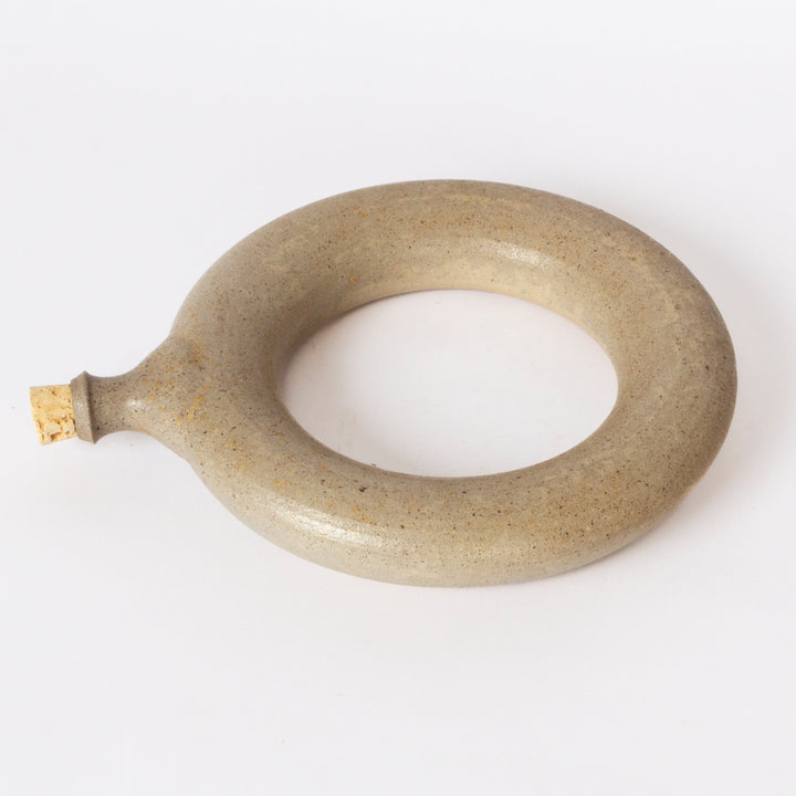 A handmade ceramic ring-shaped object with a small protruding handle, finished in a speckled light brown glaze, on a plain white background.