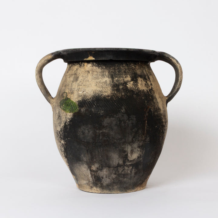 An antique ceramic double-handled jar with a distressed, weathered finish. The body is primarily black with a heavily weathered, textured beige and gray patina, featuring a small green accent on the side.