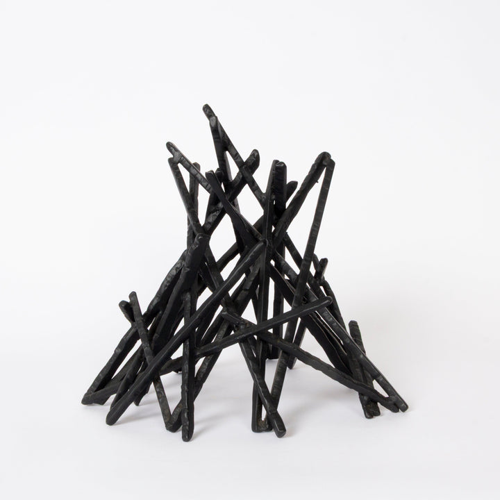 A black twig-style decorative sculpture or miniature campfire structure made of many thin intersecting sticks, photographed on a clean white studio background.