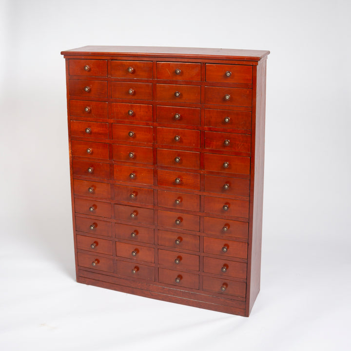 A tall wooden chest of drawers with multiple small drawers arranged in columns, featuring a warm reddish-brown finish and round metal knobs, photographed against a plain white studio background.
