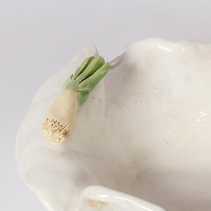 Close-up of a white ceramic dish or bowl with a sculpted onion detail featuring green tops and a cream-colored bulb, set against a plain light background.