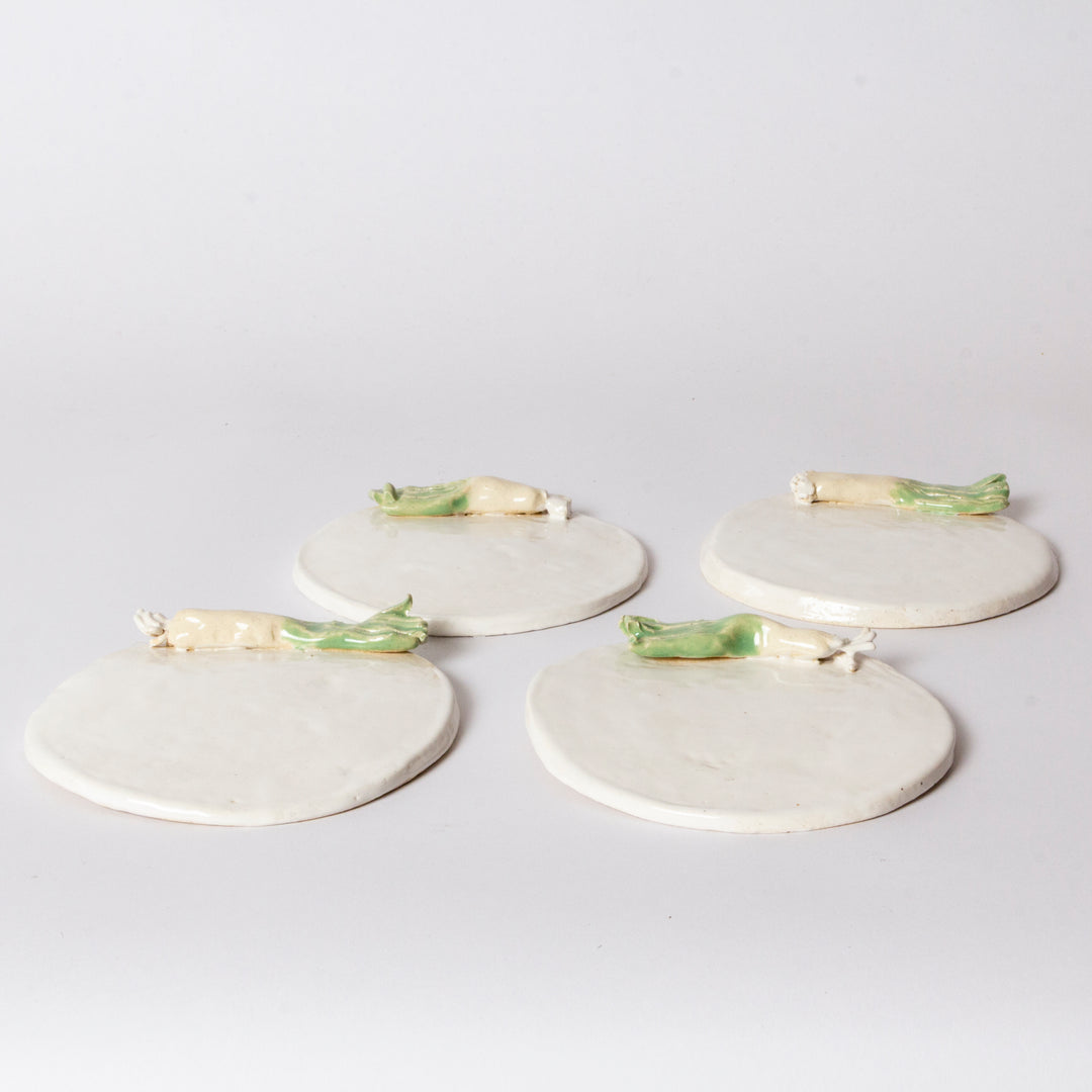 Four round off-white ceramic coasters with small green and cream vegetable figurines resting on top, arranged on a light gray background.