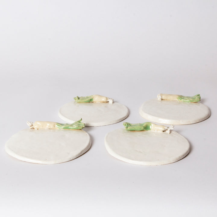 Four round off-white ceramic coasters with small green and cream vegetable figurines resting on top, arranged on a light gray background.