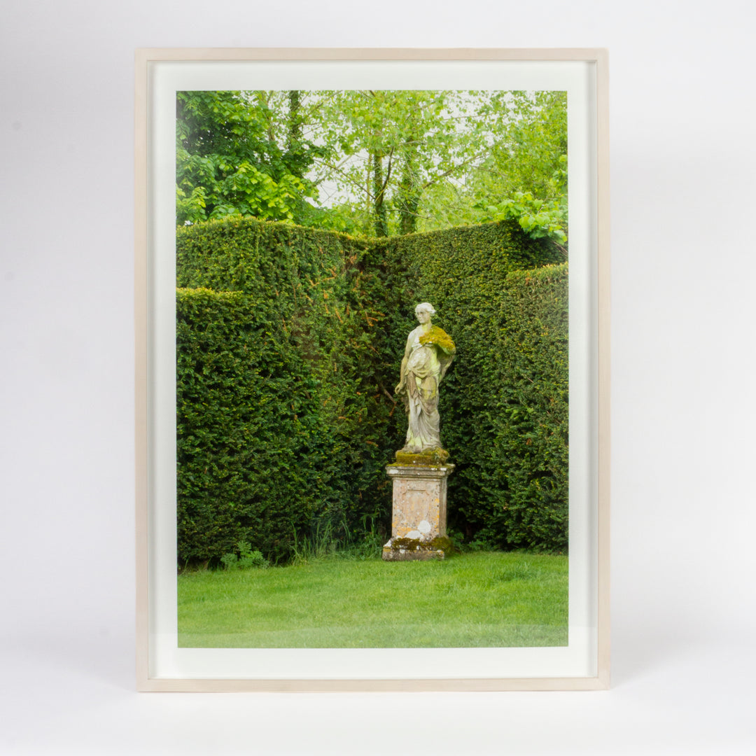 A framed art print featuring a garden scene with a mossy green hedge, a stone statue on a pedestal, and lush green grass and trees, set in a light-colored wooden frame against a white background.