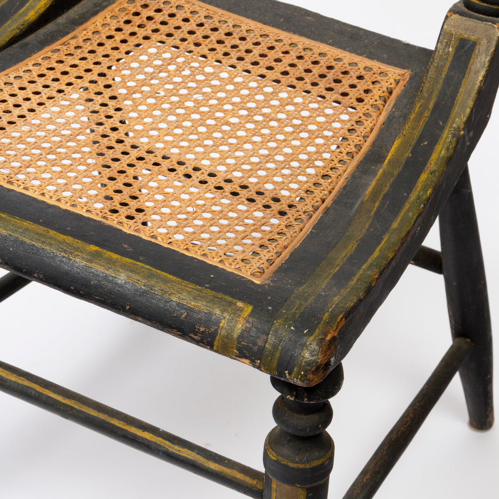 Close-up of a vintage wooden chair with a black painted frame and a natural tan woven cane seat, showing weathered gold-toned paint details.